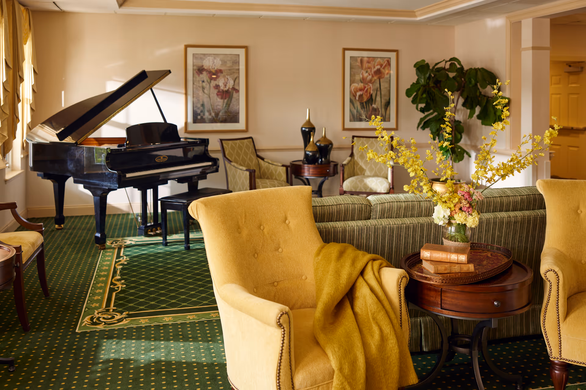 A cozy living room area with a green patterned carpet, a black grand piano near the window, two yellow armchairs with a yellow throw blanket on one, a round wooden side table with a floral arrangement and books, a striped green sofa, two framed floral paintings on the wall, and a large leafy plant in the corner.