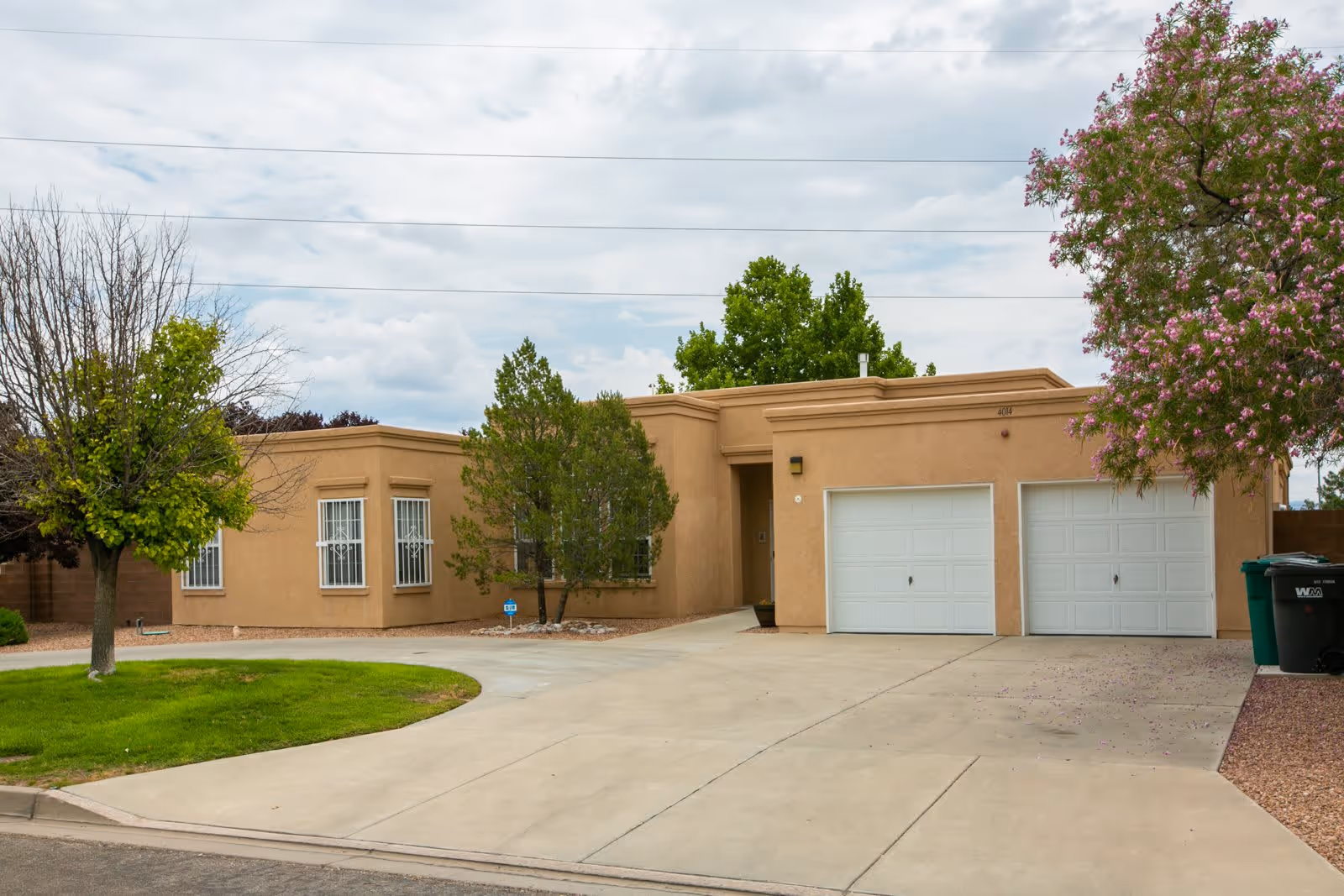 Exterior view of a single-story tan stucco building with a double garage door, a driveway, and several trees including one with pink flowers on the right side.