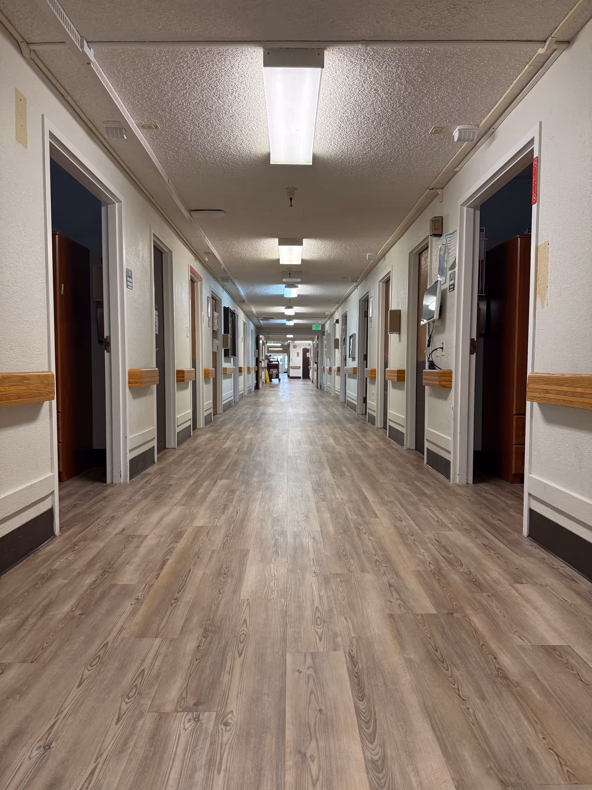 Long, empty hallway in a care facility with doors on both sides, wooden handrails, and overhead fluorescent lights.