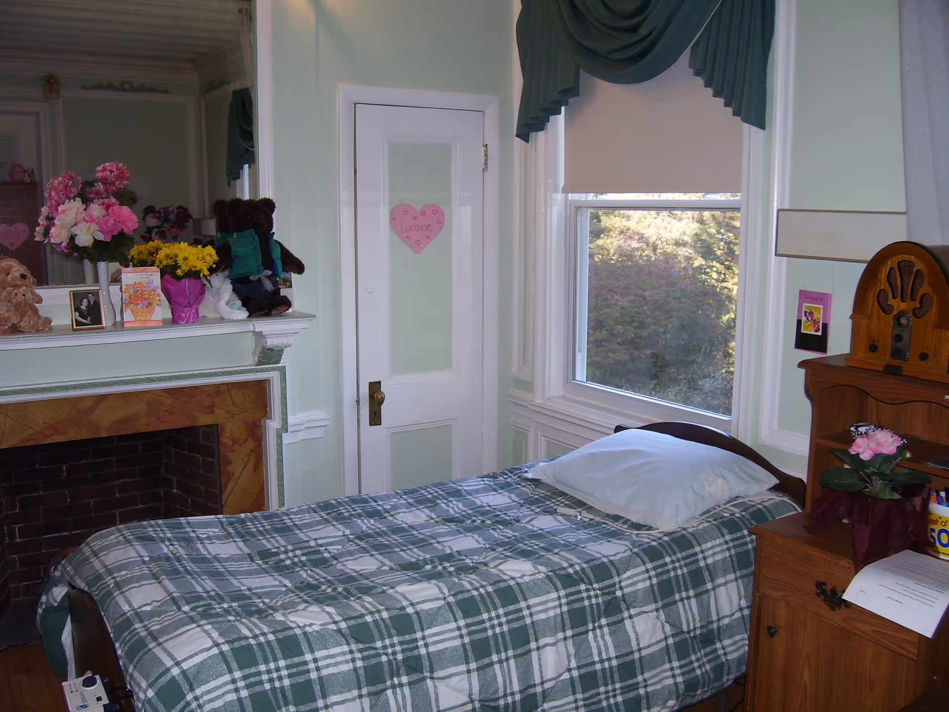 A cozy bedroom with a single bed covered in a green and white plaid blanket. There is a pillow on the bed and a wooden nightstand next to it with a pink flower pot and some papers. A vintage radio sits on a wooden shelf above the nightstand. The room has a large window with a green valance and a white roller shade, letting in natural light and showing trees outside. A white door with a pink heart-shaped note is visible, and a fireplace with a mantel decorated with flowers, teddy bears, and framed photos is on the left side.