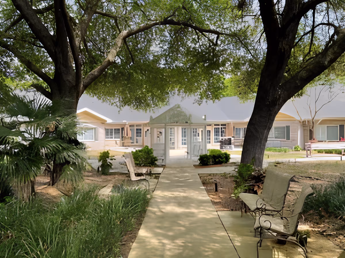 A peaceful outdoor garden area at Charter Senior Living of Gainesville featuring a paved walkway flanked by benches and lush greenery. Large trees provide shade over the path, and a building with multiple windows is visible in the background.