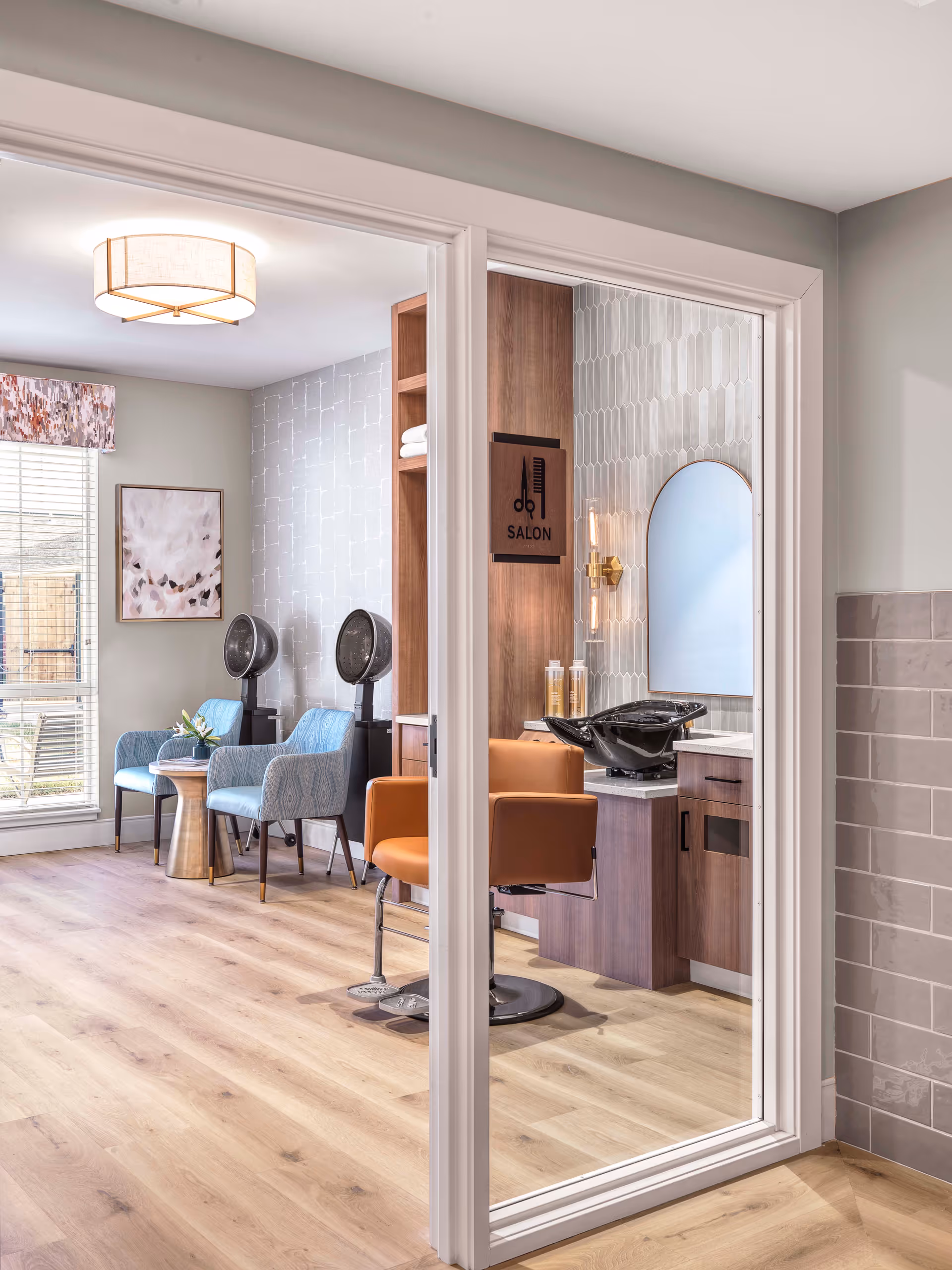 Interior view of a senior living facility's salon area with two blue chairs and hair dryers on the left side, and a salon chair with a hair washing station and mirror on the right side. The room has light wood flooring, soft lighting, and a window with blinds and a floral valance.