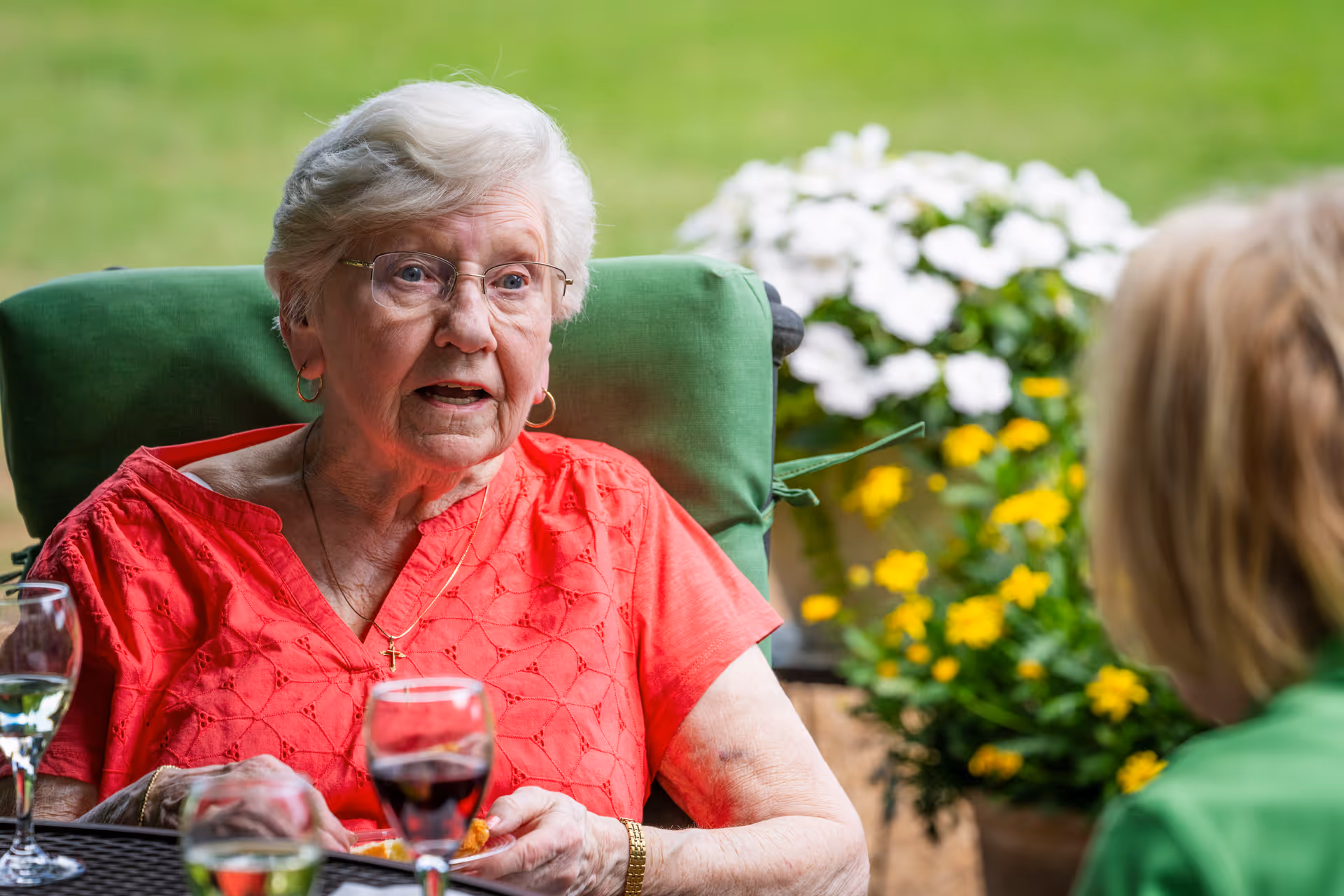 An elderly woman wearing glasses and a red blouse is sitting outdoors on a green cushioned chair, engaged in conversation with another person whose back is to the camera. There are colorful flowers in the background and glasses of wine on the table in front of them.