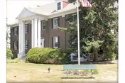 Brick two-story building with white columns at the entrance, a flagpole, bench, and landscaping in front.