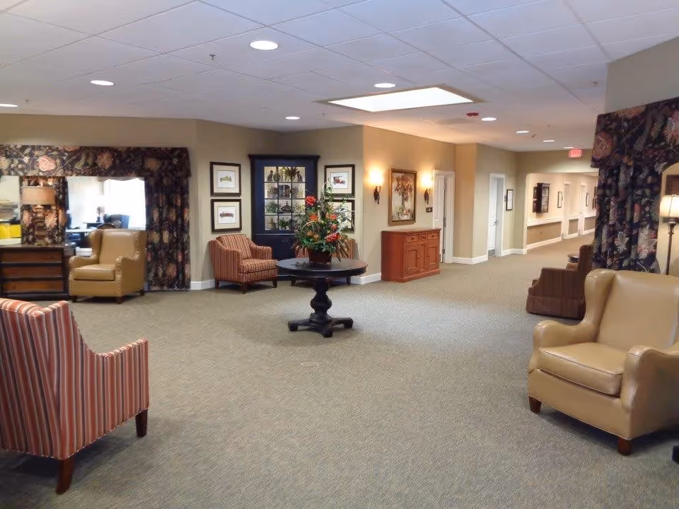 A spacious and well-lit common area in an assisted living facility featuring comfortable armchairs, a round table with a floral arrangement, framed artwork on the walls, and a hallway leading to other rooms.