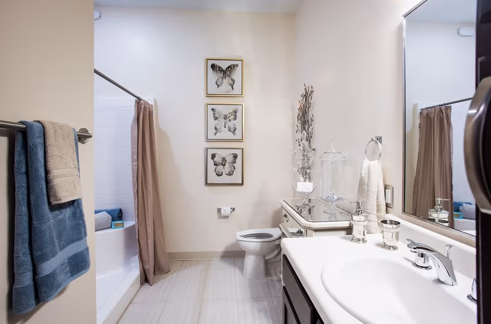 A clean and modern bathroom featuring a white sink with a silver faucet, a large mirror, a toilet, and a bathtub with a beige shower curtain. There are three framed butterfly artworks on the wall above the toilet, a towel rack with blue and beige towels, and decorative glass jars on a small cabinet next to the sink.