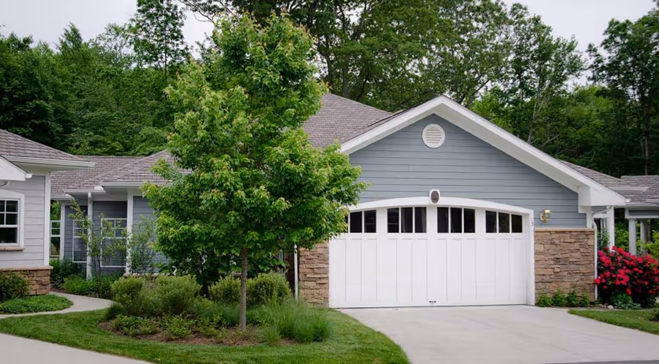 Exterior view of a residential building with a white garage door, gray siding, stone accents, and surrounded by green trees and shrubs.