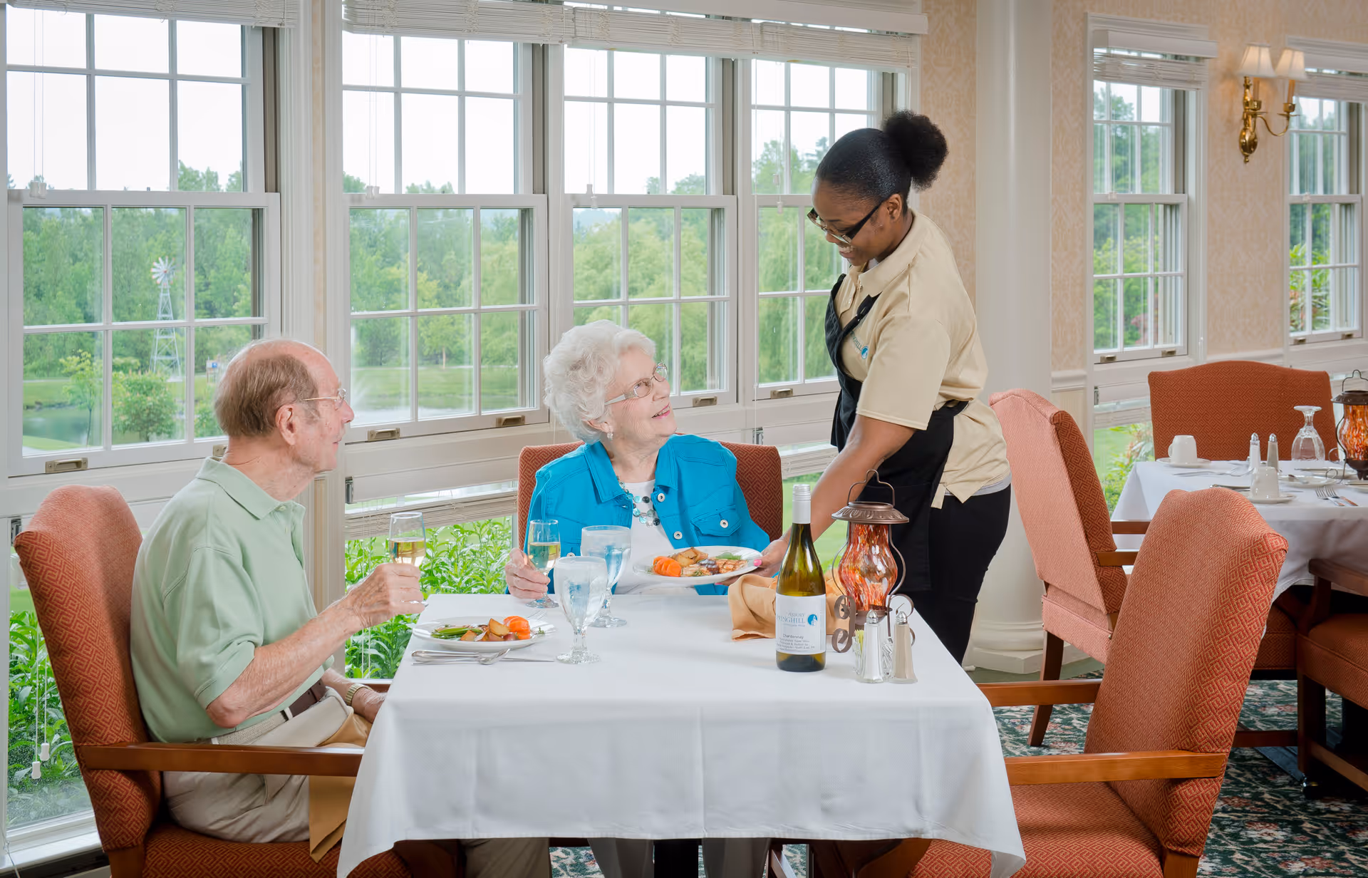 An elderly man and woman seated at a dining table in a bright room with large windows overlooking greenery. A staff member is serving a plate of food to the elderly woman. The table is set with glasses, a bottle of wine, and a decorative lantern.
