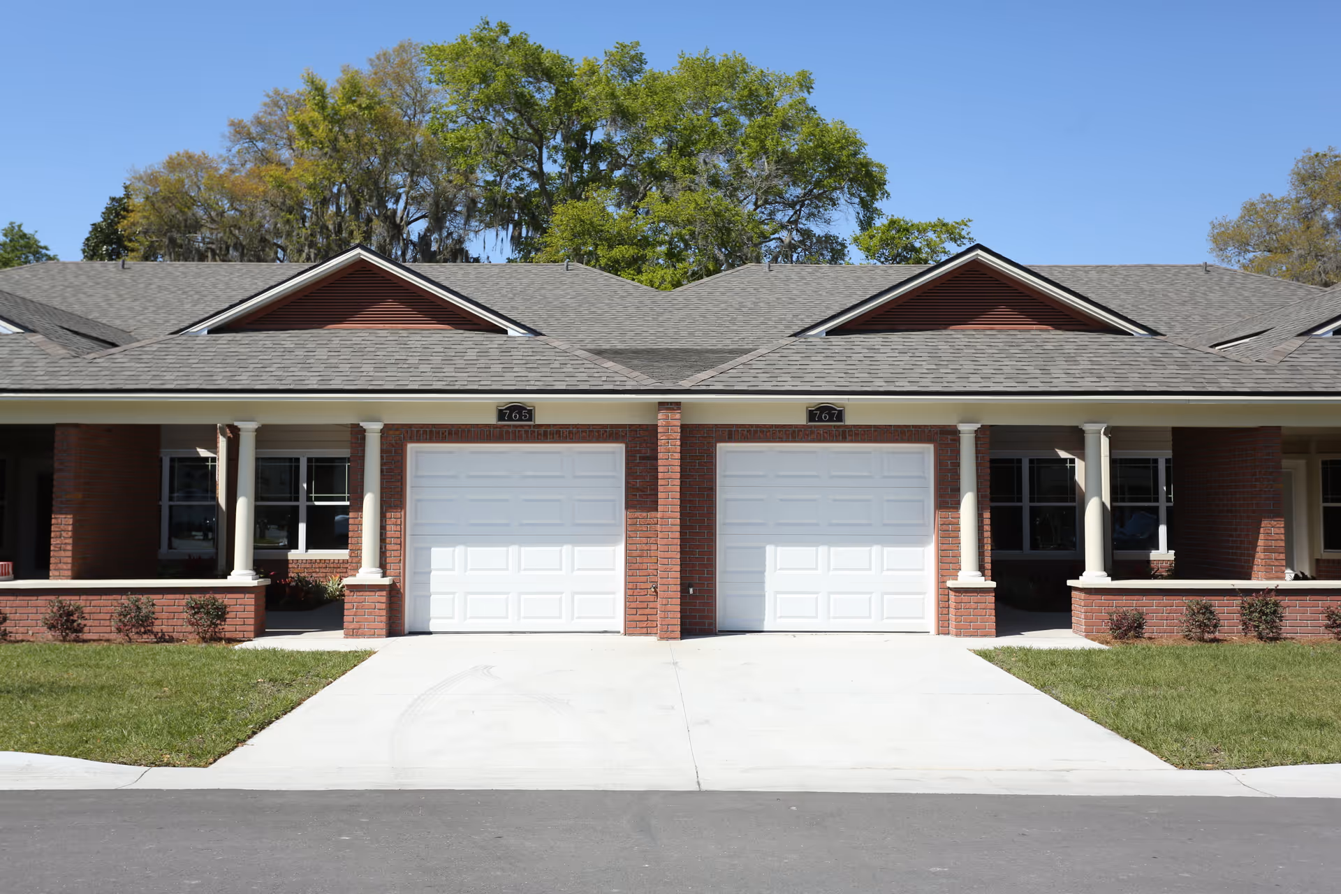 Front exterior of a single-story brick retirement duplex with two white garage doors, covered porches with columns, and a driveway.