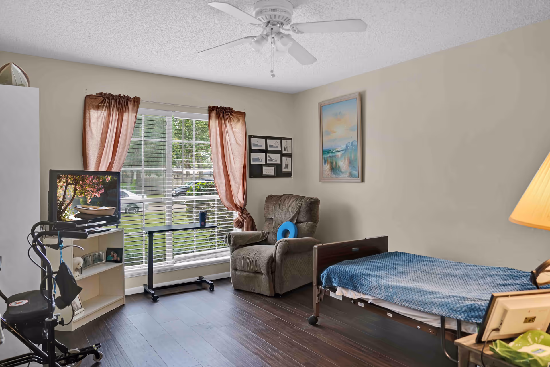 A cozy bedroom in a senior living facility with a single bed covered in a blue blanket, a comfortable recliner chair with a blue cushion, a small TV on a white shelf, a rolling tray table, and a large window with brown curtains letting in natural light. The room has wooden flooring, beige walls with framed artwork, and a ceiling fan.