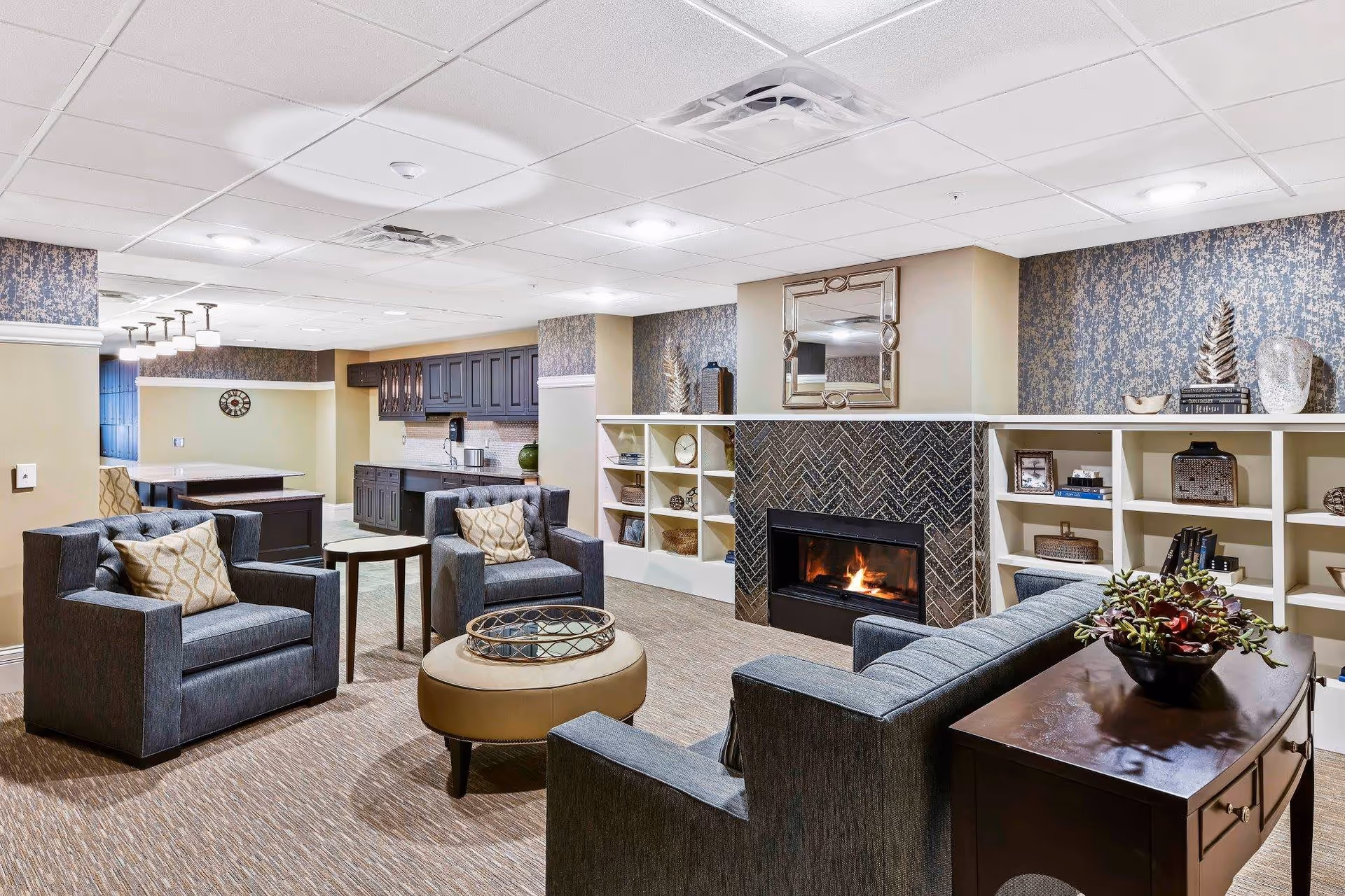 A cozy living room area in a senior living facility with a fireplace surrounded by a herringbone tile pattern. The room features four gray upholstered armchairs with patterned pillows arranged around a round ottoman with a decorative tray on top. Behind the fireplace are built-in shelves with decorative items and books. The walls have a blue and beige patterned wallpaper, and there is a large mirror above the fireplace. In the background, there is a kitchenette area with dark cabinets and a dining table with chairs.