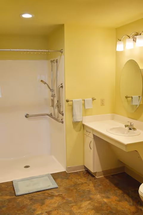 A bathroom with a walk-in shower featuring grab bars and a handheld showerhead. There is a towel rack with two white towels, a sink with a countertop, an oval mirror above the sink, and a three-light fixture on the wall. The walls are painted yellow and the floor has a brown patterned finish.