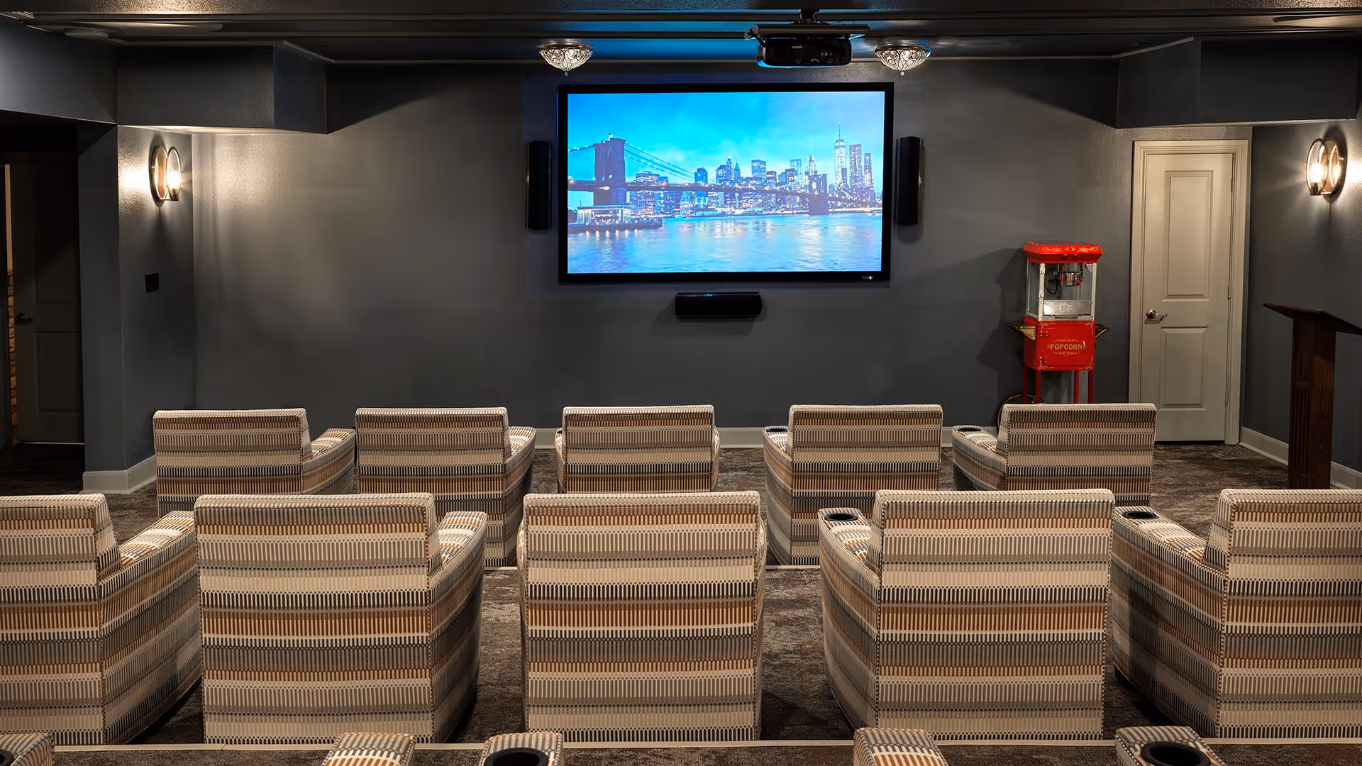 A small home theater room with two rows of striped armchairs facing a large flat-screen TV mounted on a dark gray wall. A red popcorn machine is positioned near a closed white door on the right side of the room. The room has dim wall sconces and a carpeted floor.