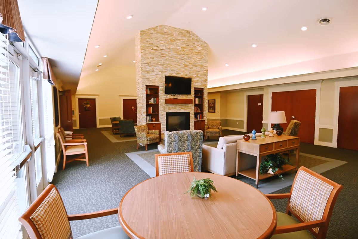 A spacious and well-lit common living area in The Cortland Wyoming featuring a round wooden table with four chairs in the foreground, a stone fireplace with a mounted TV above it, and several upholstered chairs and sofas arranged around the fireplace. Large windows with blinds allow natural light to fill the room, and there are wooden doors and decorative elements along the walls.