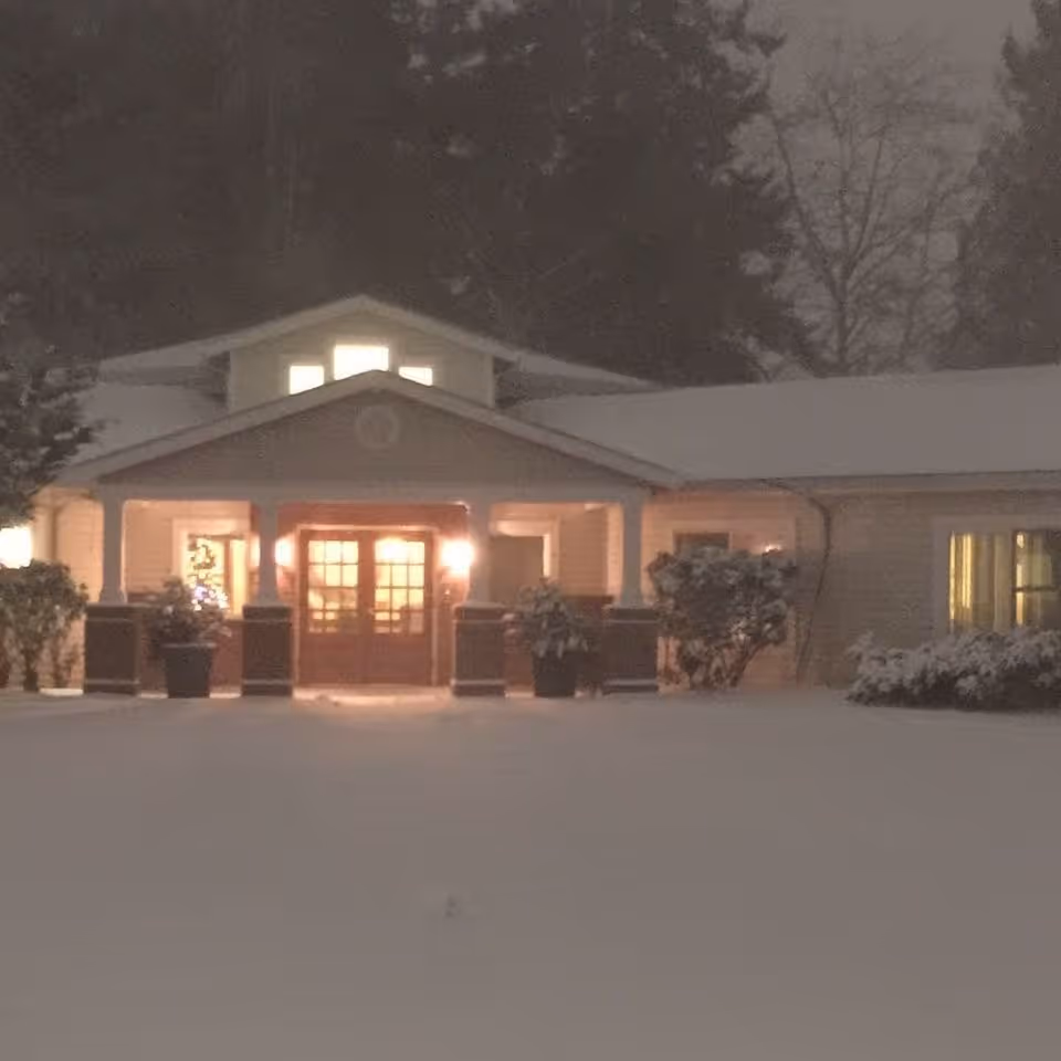Snow-covered front entrance of a low building with a lit porch and windows at night.