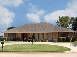 Single-story residential building with a wide, low-pitched roof, multiple windows with red shutters, a central front door, and neatly trimmed bushes along the front. The house is set behind a well-maintained lawn and a curved driveway under a partly cloudy sky.