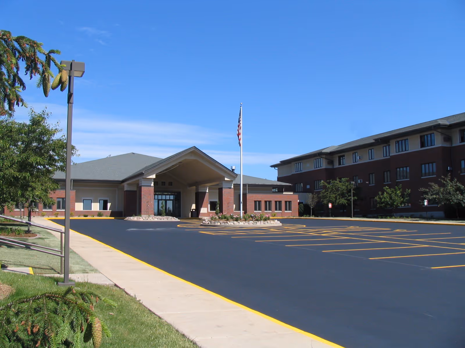 Exterior view of a senior living facility with a large parking lot in front, a flagpole with an American flag, and a clear blue sky. The building has a covered entrance with brick and beige walls, and there are trees and landscaping around the property.