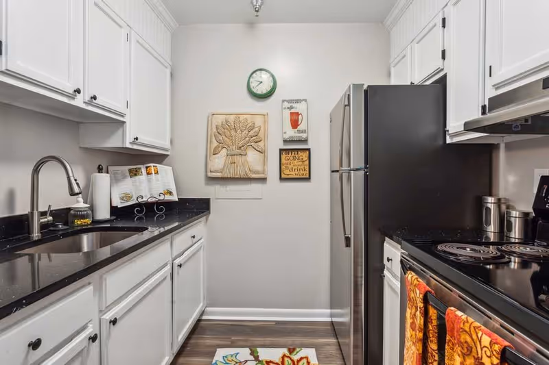 A modern kitchen with white cabinets, black countertops, a stainless steel refrigerator, and an electric stove with colorful towels hanging on the handle. There is a sink with a faucet on the left side, a cookbook on a stand, and three decorative wall hangings including a clock and coffee-themed signs on the far wall. The floor has a colorful floral rug.