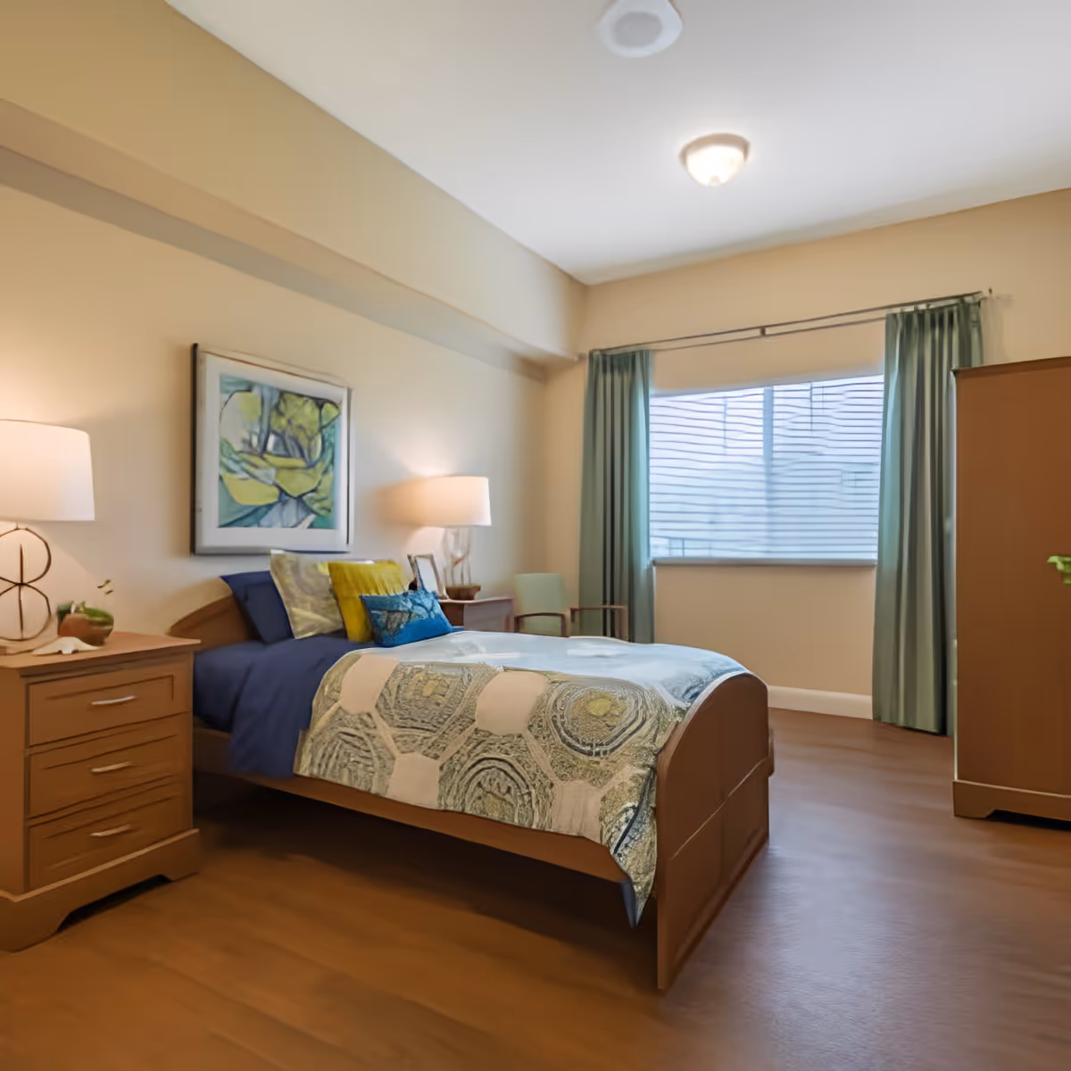 Furnished senior living bedroom with a single bed, nightstand and lamps, artwork on the wall, and a window with green curtains.