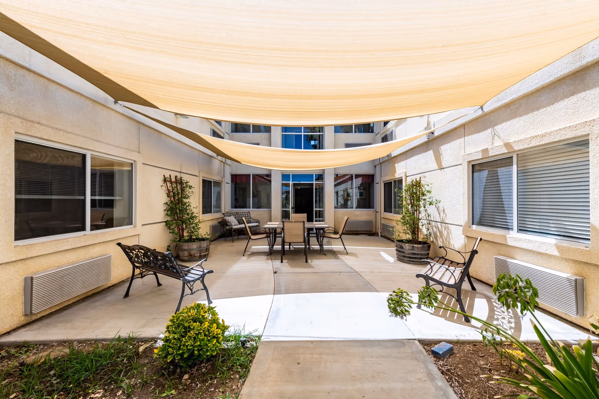 Outdoor courtyard area with beige shade sails overhead, two black metal benches on either side, potted plants, and a table with chairs in the center. The courtyard is surrounded by beige walls with windows and a door at the far end.