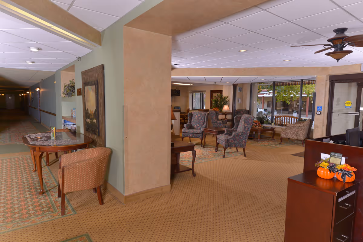 Senior living lobby with armchairs, tables, a reception desk, and a front entrance with large windows.
