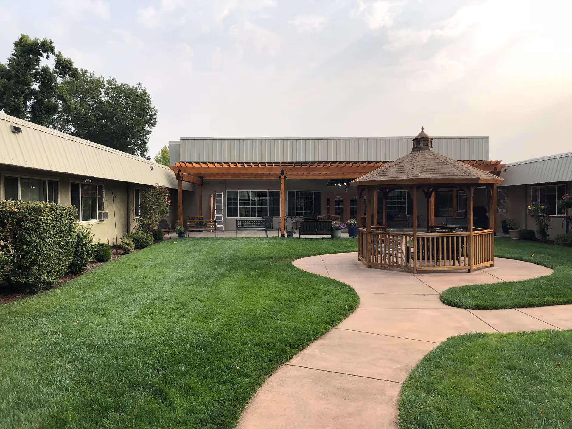 Outdoor courtyard area of a senior living facility with a wooden gazebo in the center, surrounded by a curved concrete pathway and green grass. The building surrounds the courtyard with windows and a covered patio area with benches and potted plants.