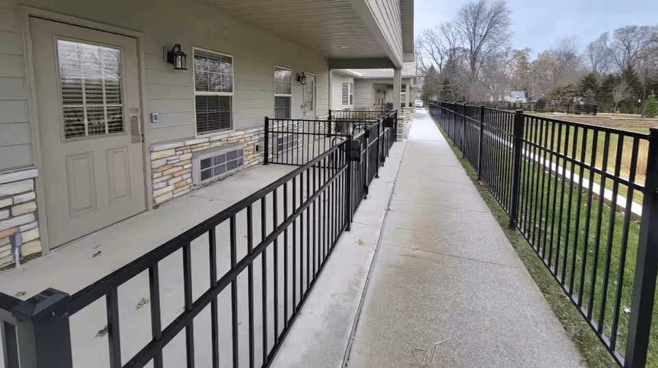 Outdoor walkway alongside a building with beige siding and stone accents, featuring multiple doors and windows. Black metal railings separate the walkway from a grassy area on the right. Leafless trees and a clear sky are visible in the background.