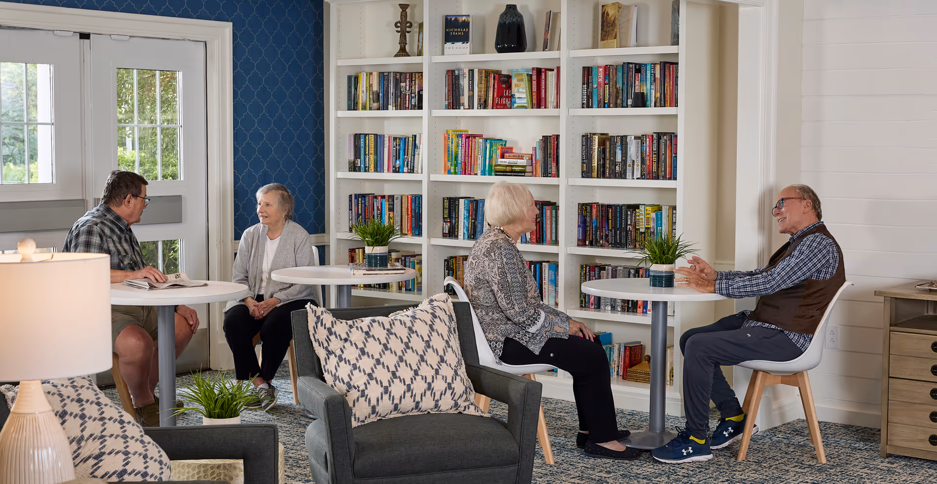 A cozy common area in Monarch Spring Meadows with two small round tables where four elderly people are seated and engaged in conversation. The room features a large white bookshelf filled with books, blue patterned wallpaper on one wall, a lamp on a side table, and comfortable armchairs with patterned cushions.