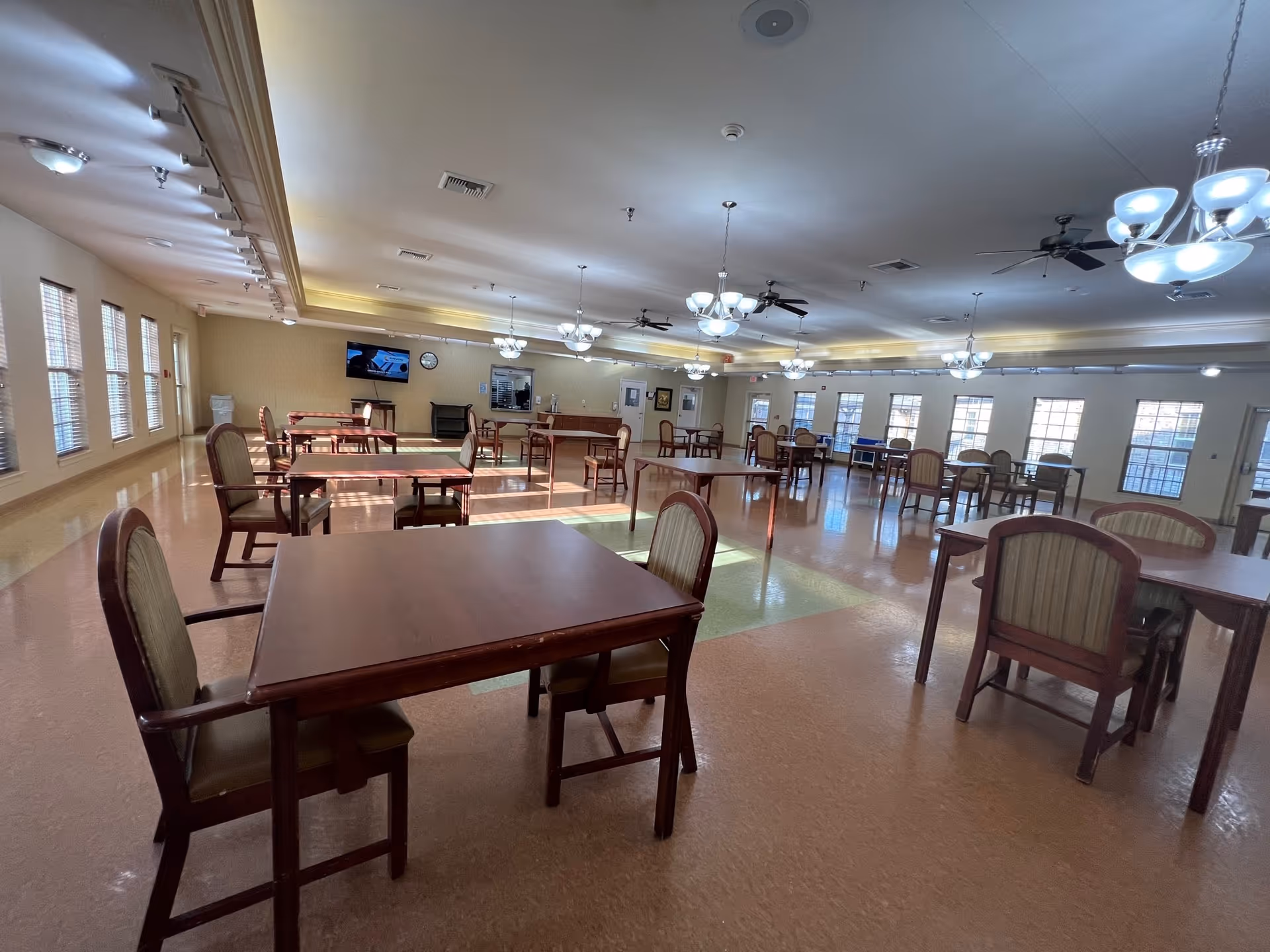 A spacious dining room with multiple wooden tables and chairs arranged neatly. The room has large windows allowing natural light to fill the space, ceiling fans, and hanging light fixtures. A television and a clock are mounted on the far wall.