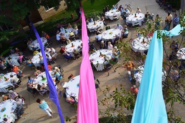 Aerial view of an outdoor gathering at Sentinel Pointe Retirement Community with multiple round tables covered with white tablecloths. People are seated around the tables enjoying the event. Colorful fabric banners in purple, pink, and light blue hang vertically in the foreground. Trees and greenery surround the paved area.