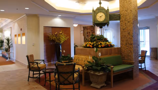 Interior common area with seating including cushioned chairs and a green bench surrounding a central planter with flowers and a vintage-style clock above it. The space features tiled floors, potted plants, and soft lighting with a warm color palette.