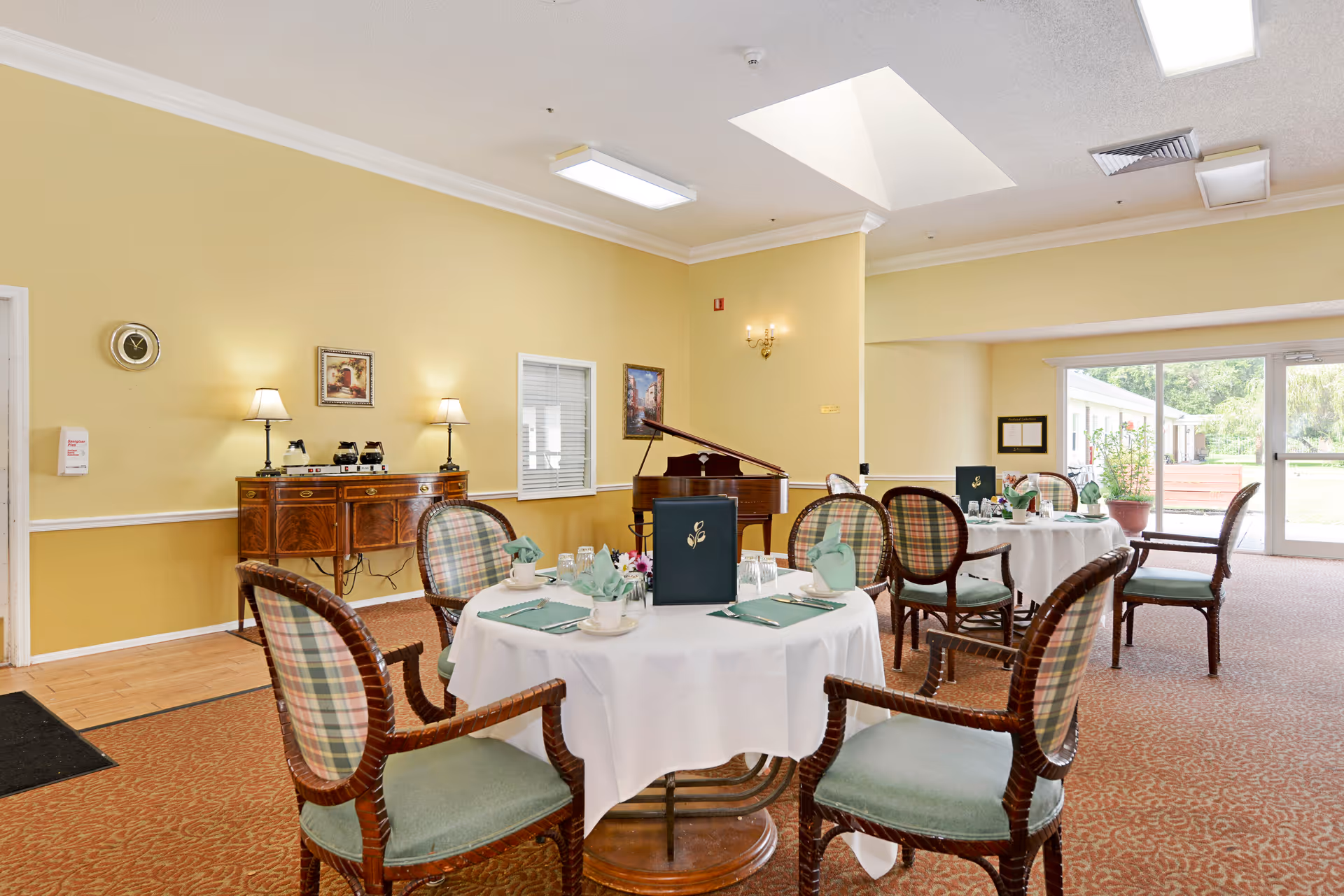 Dining area in Brookdale Colonial Park with round tables covered in white tablecloths, set with green napkins, cups, and glasses. The room has yellow walls, plaid cushioned chairs, a wooden sideboard with lamps and coffee pots, a grand piano in the corner, and large windows letting in natural light.