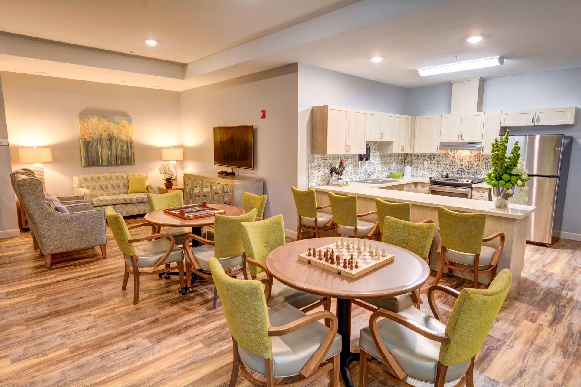 A bright and inviting common area in a memory care facility featuring a kitchen with light wood cabinets, a stainless steel refrigerator, and a patterned tile backsplash. In front of the kitchen is a counter with six green upholstered chairs. There are two round tables with green chairs around them, one table has a chessboard set up. In the background, there is a living room area with a sofa, armchairs, two lamps, a wall-mounted TV, and a decorative painting on the wall. The floor is wood-style laminate.