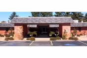 Front entrance of a one-story brick nursing and rehab building with a covered drive-up portico, parking spaces, and low shrubs.