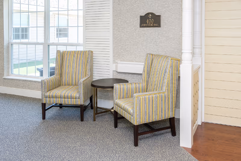 Two striped armchairs with yellow, blue, and gray colors are positioned around a small round wooden table in a well-lit room with a large window and light-colored walls. A sign on the wall reads '400 CONVENTION HALL'.