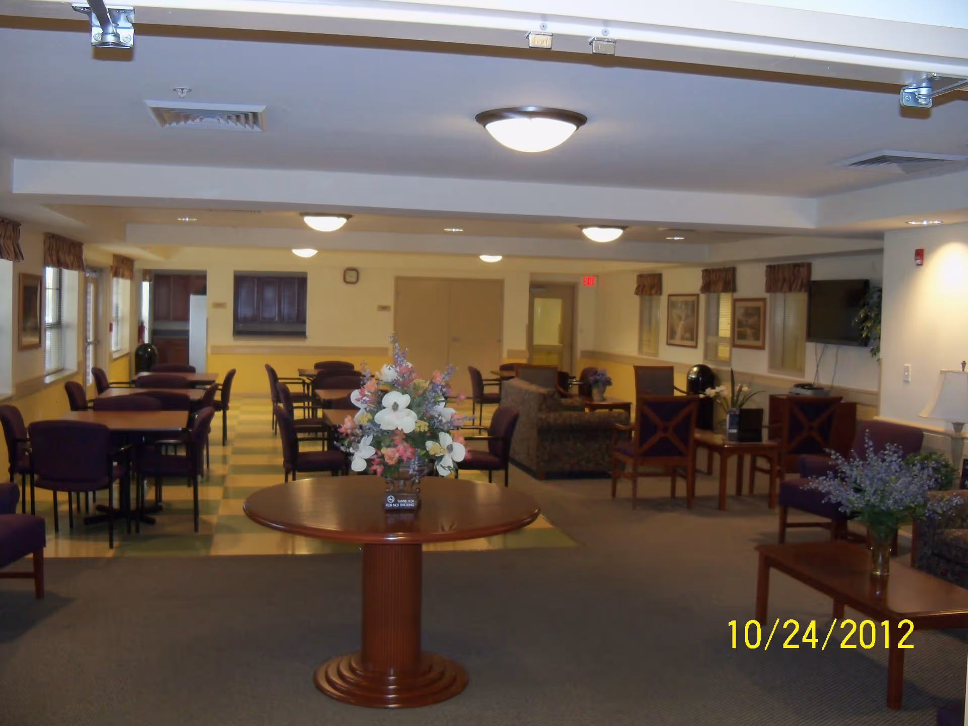 Interior view of a senior living facility common area with multiple tables and chairs arranged for dining and socializing. There is a round wooden table with a floral arrangement in the foreground, several square tables with purple chairs in the background, and a seating area with couches and armchairs near a wall-mounted TV. The room is well-lit with ceiling lights and has windows with valances along one side.