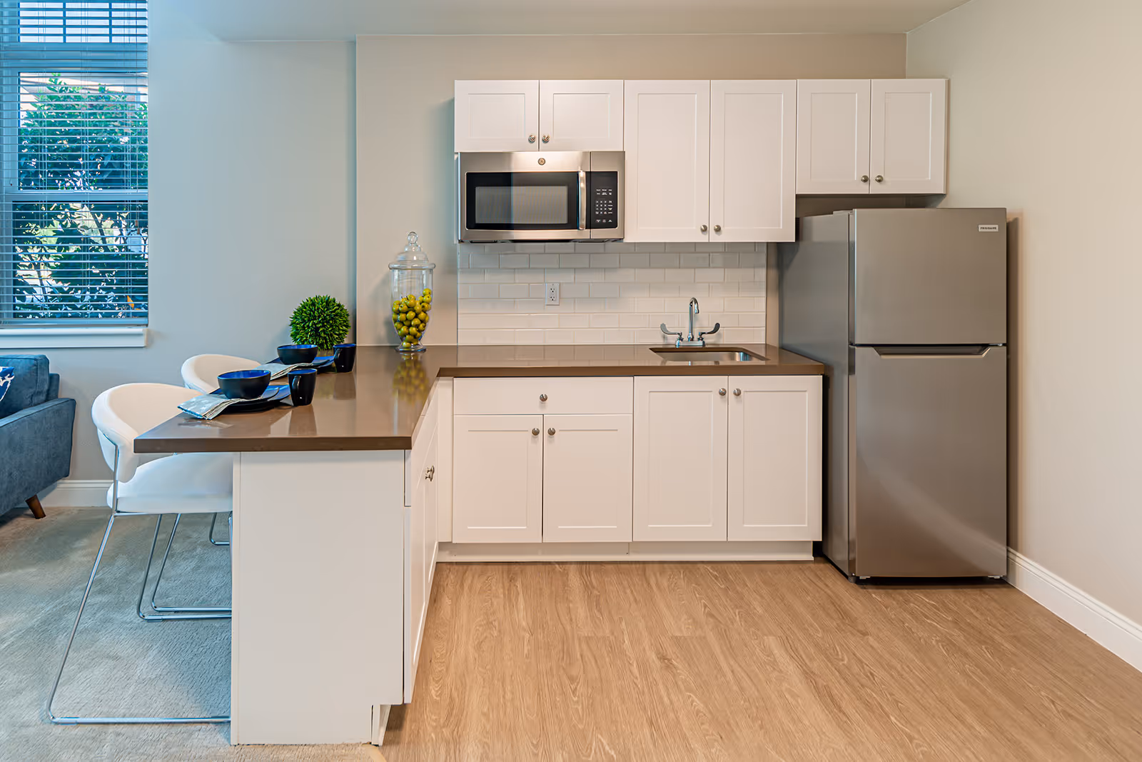 Modern kitchen area with white cabinets, a stainless steel microwave and refrigerator, a small sink, and a brown countertop. There is a glass jar filled with green apples and a small green plant on the counter. Adjacent to the kitchen is a dining area with two white chairs and a table set with blue bowls and black mugs. A window with blinds lets in natural light, and part of a gray sofa is visible in the background.
