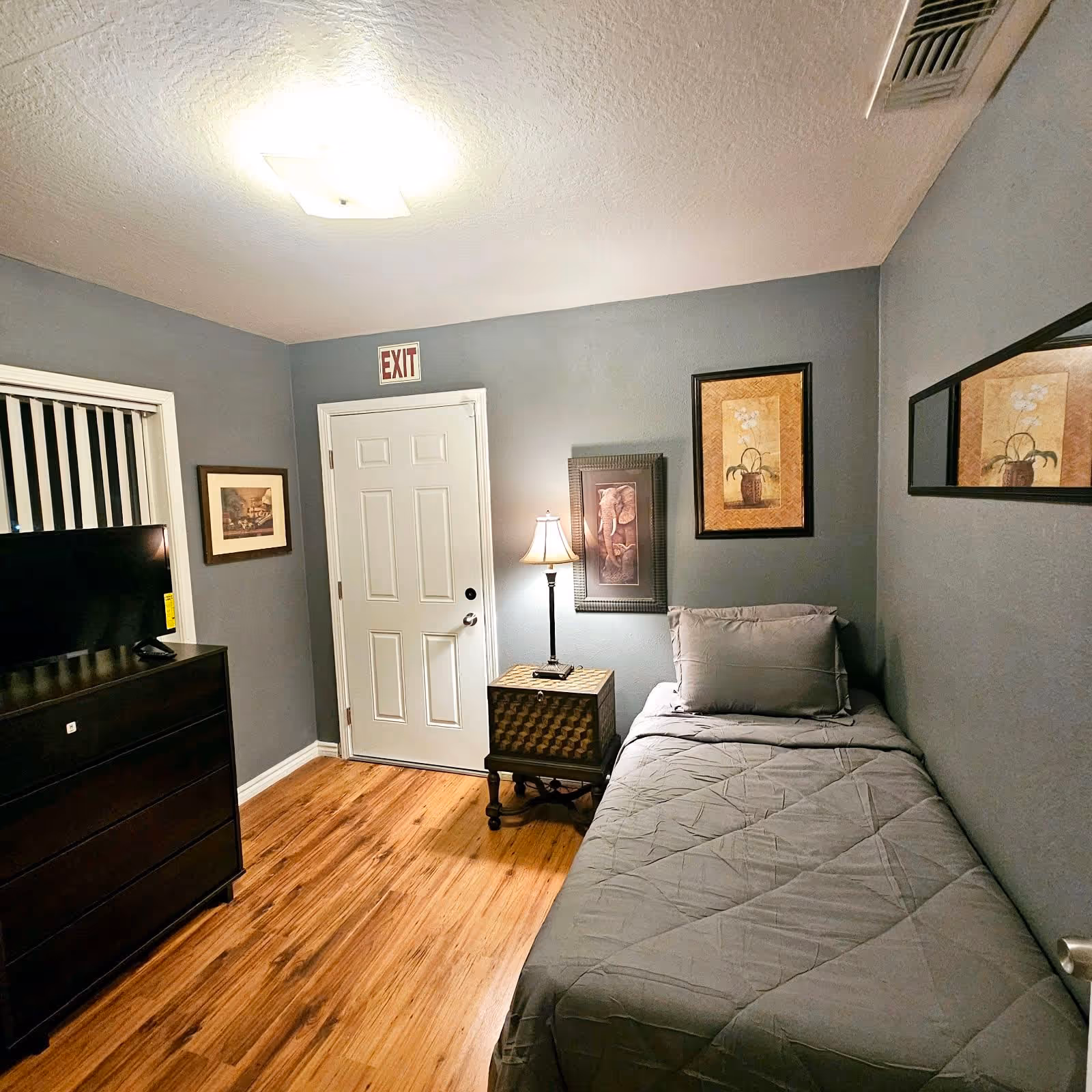 A small bedroom with gray walls and wooden flooring featuring a single bed with gray bedding and a pillow. Next to the bed is a small wooden nightstand with a lamp. On the wall above the nightstand are two framed pictures, one of an elephant and another of a flower in a basket. Across from the bed is a dark wooden dresser with a flat-screen TV on top. A white door with an exit sign above it is visible at the end of the room, and a rectangular mirror hangs on the right wall.