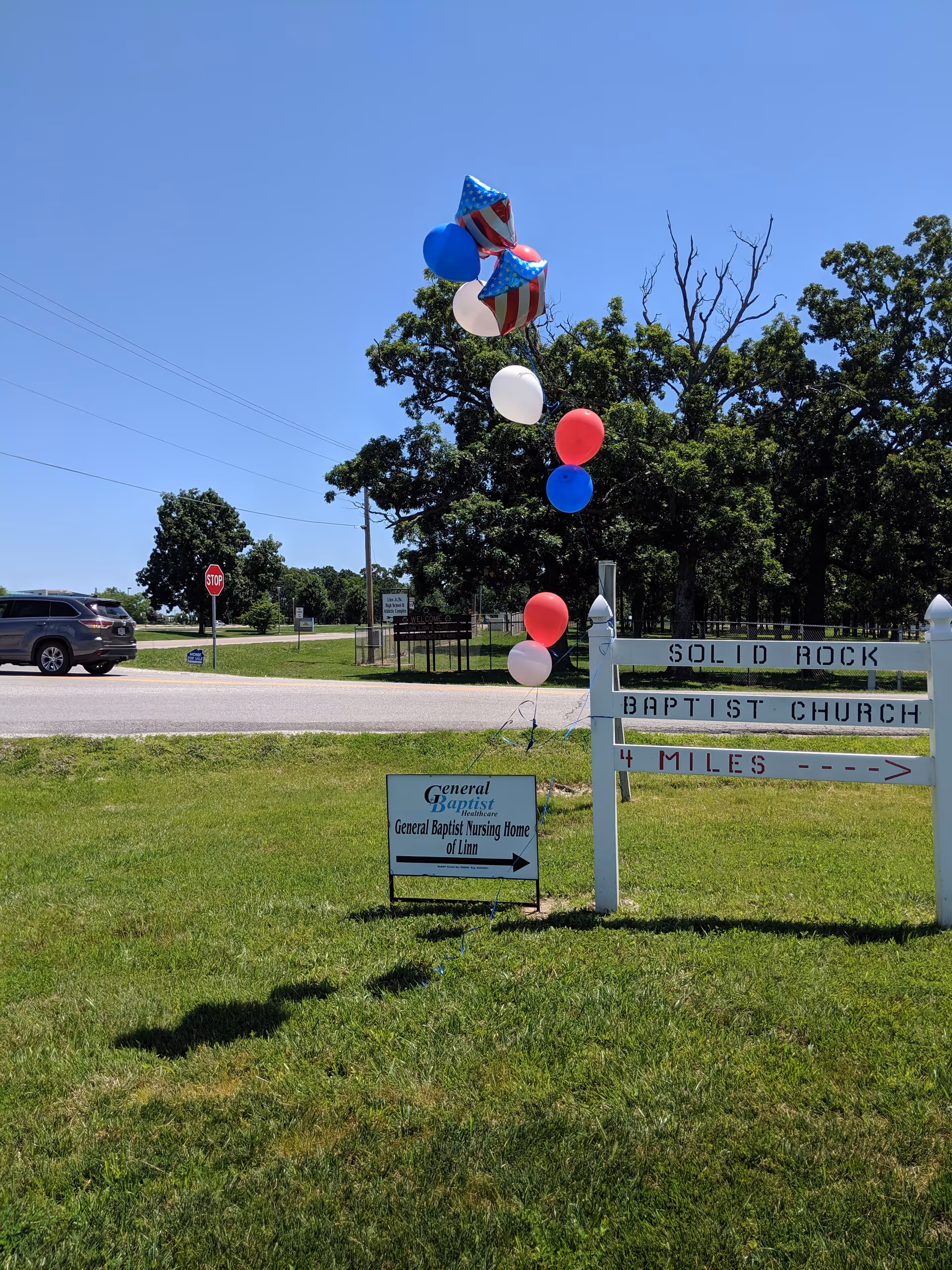 Outdoor scene showing a grassy area with a sign for General Baptist Nursing Home of Linn and a larger sign for Solid Rock Baptist Church. Red, white, and blue balloons are tied to the nursing home sign. A road, a stop sign, a parked car, and trees are visible in the background under a clear blue sky.