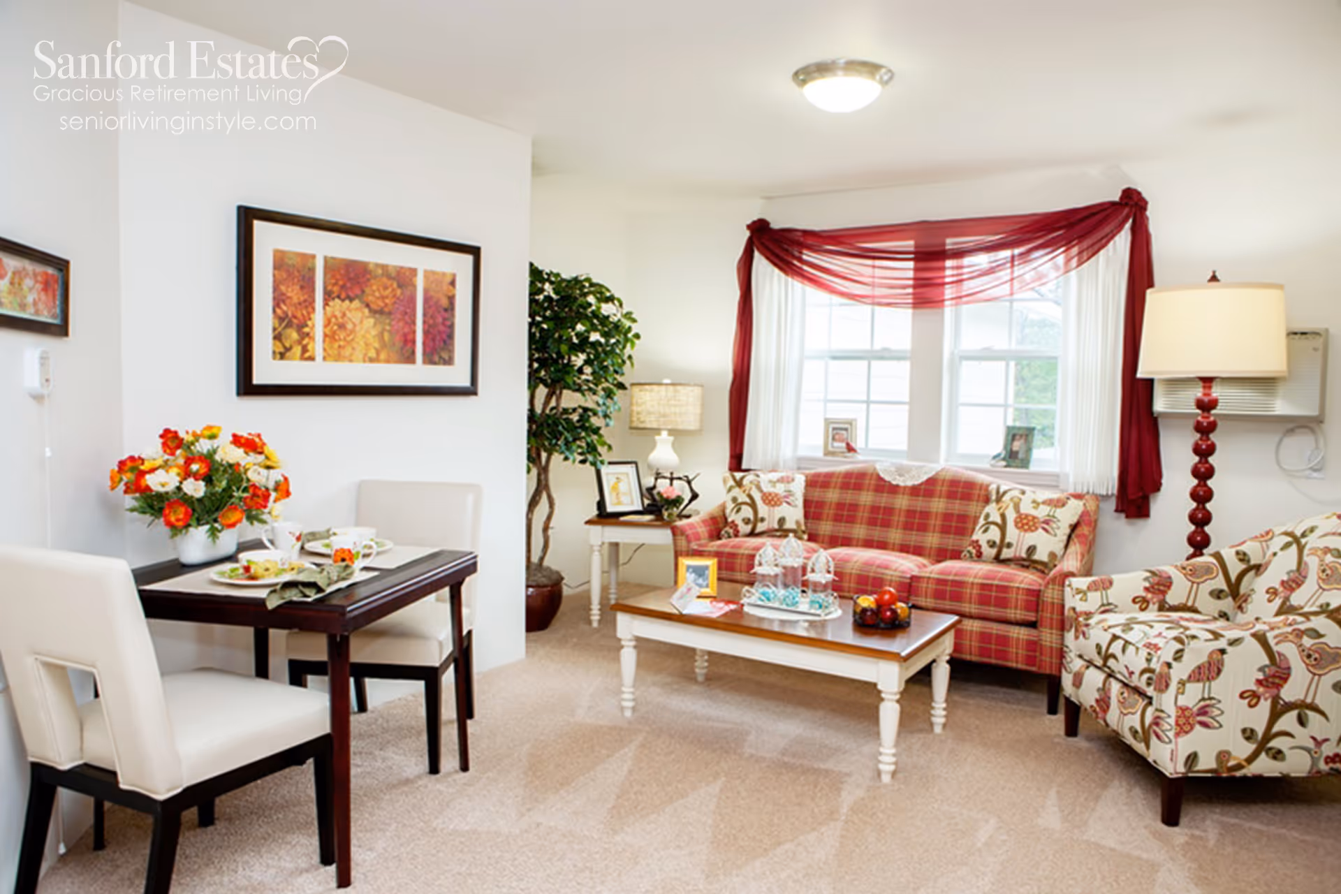A cozy living room and dining area in a retirement living facility. The living room features a red plaid sofa with patterned cushions, a floral armchair, a wooden coffee table with decorative items, a floor lamp, and a window with sheer white curtains and a red swag valance. The dining area has a small dark wooden table set for two with white cushioned chairs and a vase of colorful flowers. The walls are decorated with framed floral artwork and there is a potted plant in the corner.