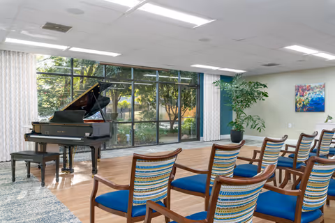 Sunlit community activity room with a grand piano and rows of upholstered chairs facing large floor-to-ceiling windows.