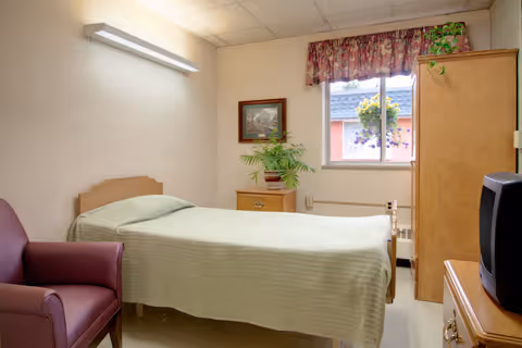 A small bedroom in a senior living facility with a single bed covered with a light green blanket, a maroon armchair, a wooden nightstand with a potted plant, a framed picture on the wall, a window with floral curtains, and a wooden wardrobe. A TV is placed on a wooden dresser in the foreground.