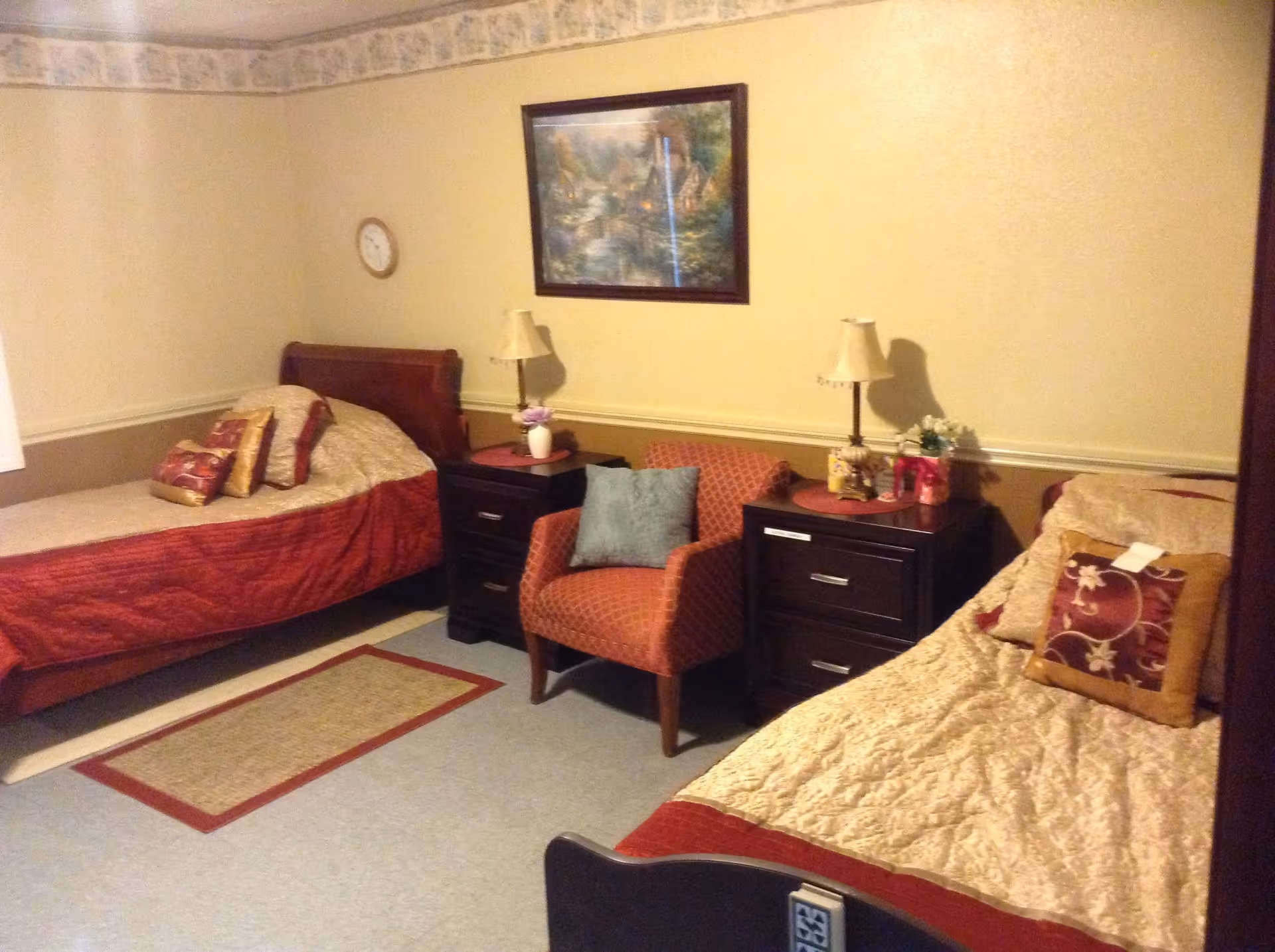 A cozy bedroom with two single beds, each with a quilted bedspread and decorative pillows. Between the beds are two dark wooden nightstands with lamps, a small vase with flowers, and other small decorative items. A patterned armchair with a blue cushion is placed between the nightstands. A framed painting hangs on the beige wall above the chair, and a small clock is mounted on the wall near one bed. The room has a light blue floor and a small rectangular rug with a red border.