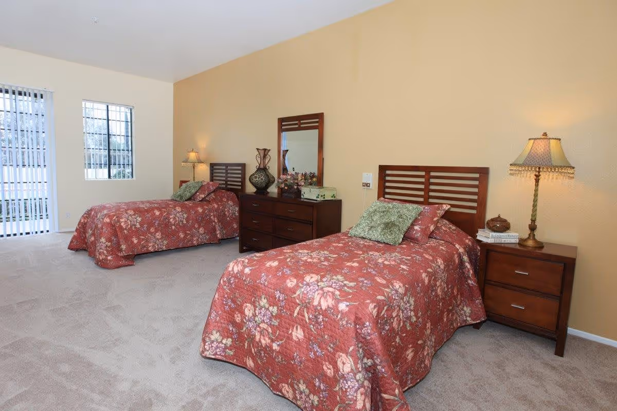 A bedroom with two twin beds covered in red floral bedspreads, each with green and red pillows. Between the beds is a wooden dresser with a mirror, decorative vase, and flowers. There is a nightstand with a lamp and books next to the bed on the right. The room has beige walls, carpeted floor, and two windows with vertical blinds letting in natural light.