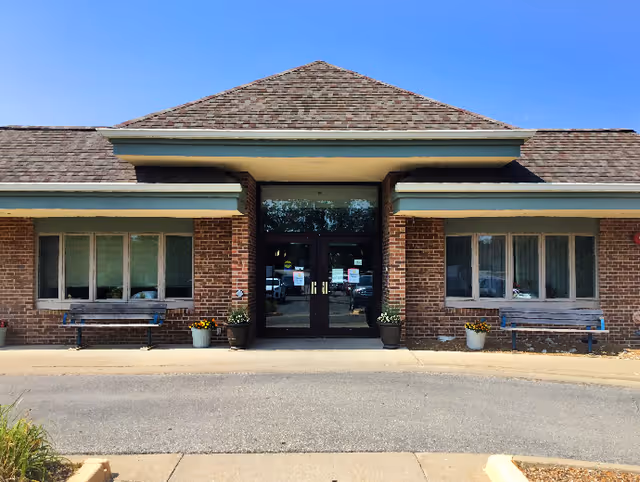 Front exterior view of a brick building with a peaked roof and double glass entrance doors. There are two benches and potted plants on either side of the entrance. The sky is clear and blue.