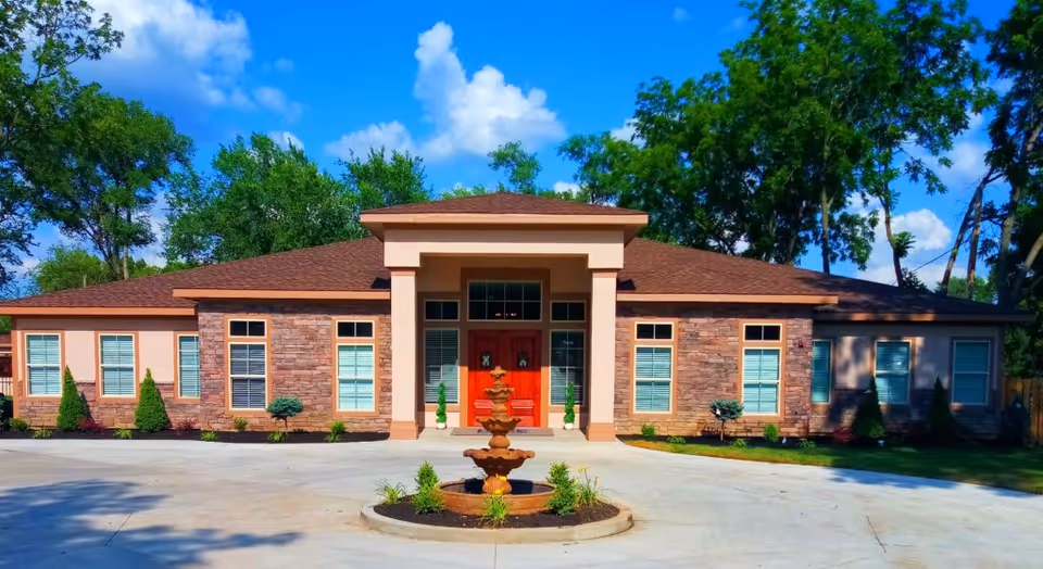 Single-story senior care building with a central entrance and red double doors, brick facade, circular driveway and a fountain in front under a blue sky.