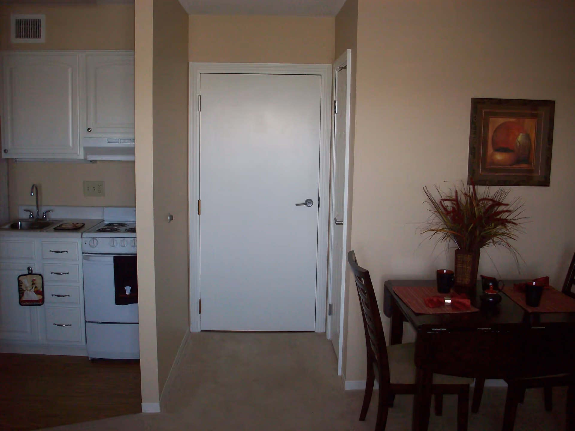 Interior view showing a small kitchen area with white cabinets, a stove, and a sink on the left, a closed white door in the center, and a dining table with chairs, placemats, cups, and a vase with decorative plants on the right. A framed artwork hangs on the wall above the dining table.