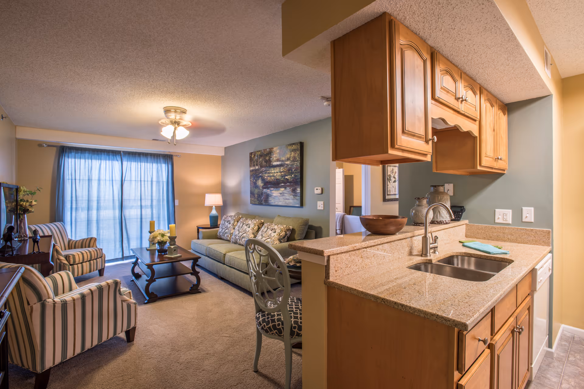 Open-plan living area with a sofa, striped armchairs, coffee table, and an adjacent kitchen counter with sink and wooden cabinets.