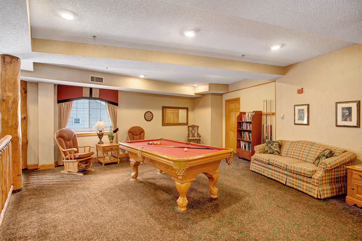 A cozy recreational room with a red felt pool table in the center, two wooden rocking chairs with cushions near a window with curtains, a small side table with a lamp and plant, a dartboard on the wall, a plaid sofa with patterned pillows, a wooden bookshelf filled with books, and framed artwork on the walls.