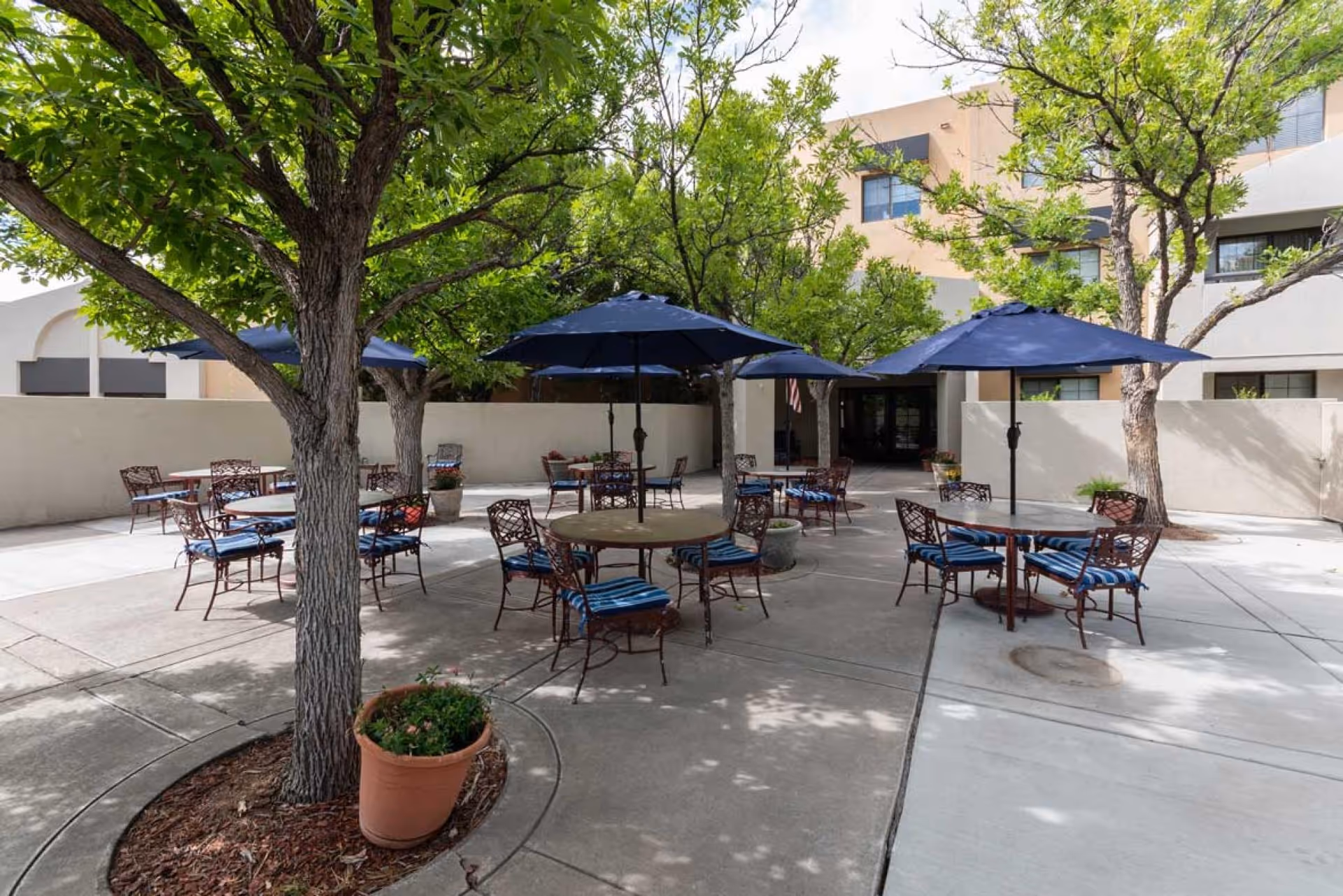 Outdoor patio area with several round tables and metal chairs with blue striped cushions. Each table has a large blue umbrella providing shade. The patio is surrounded by trees and potted plants, with a beige building in the background.